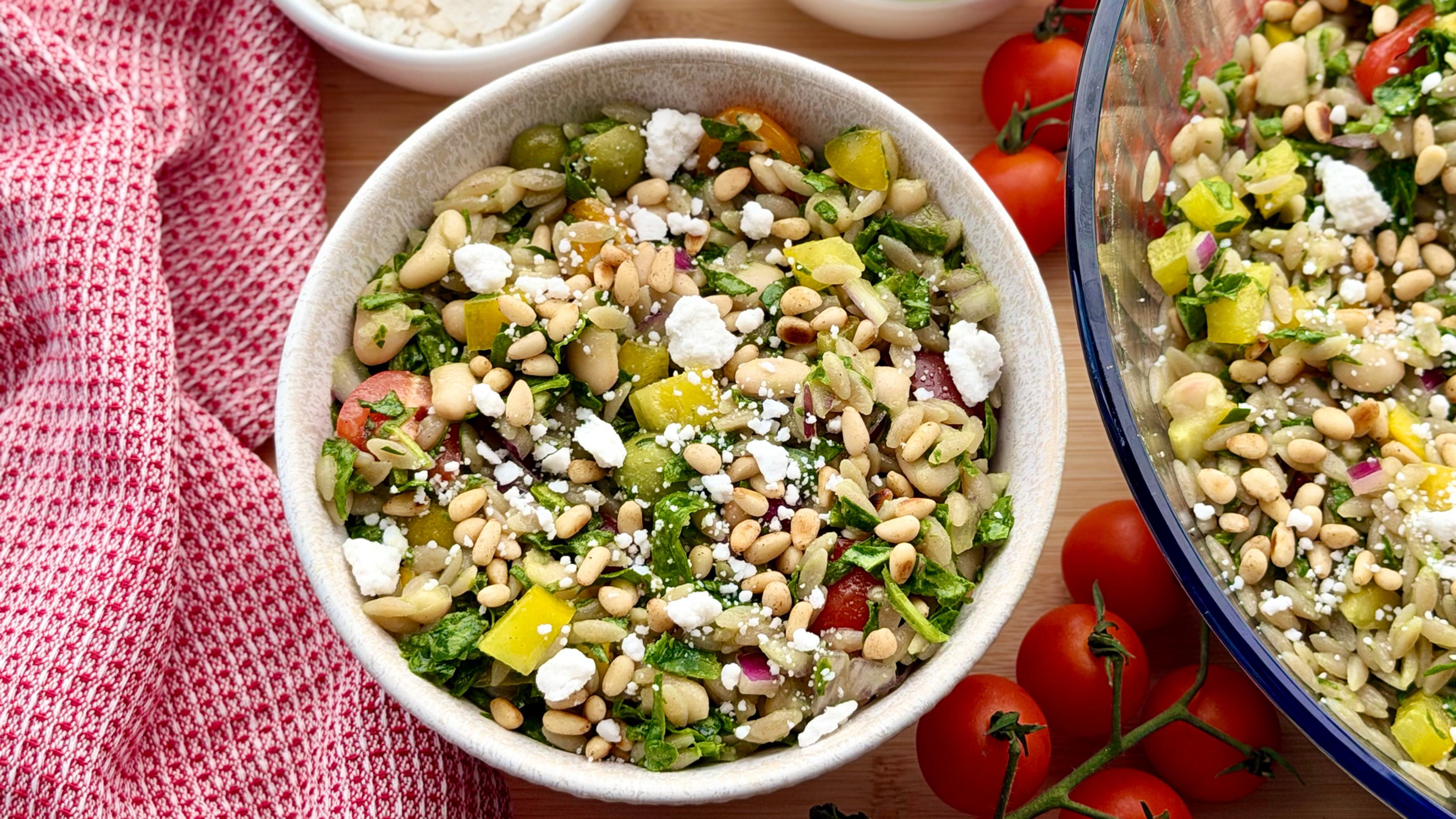 A bowl of gluten free orzo cassava pasta salad with bowls of feta and spinach and tomatoes on the vine