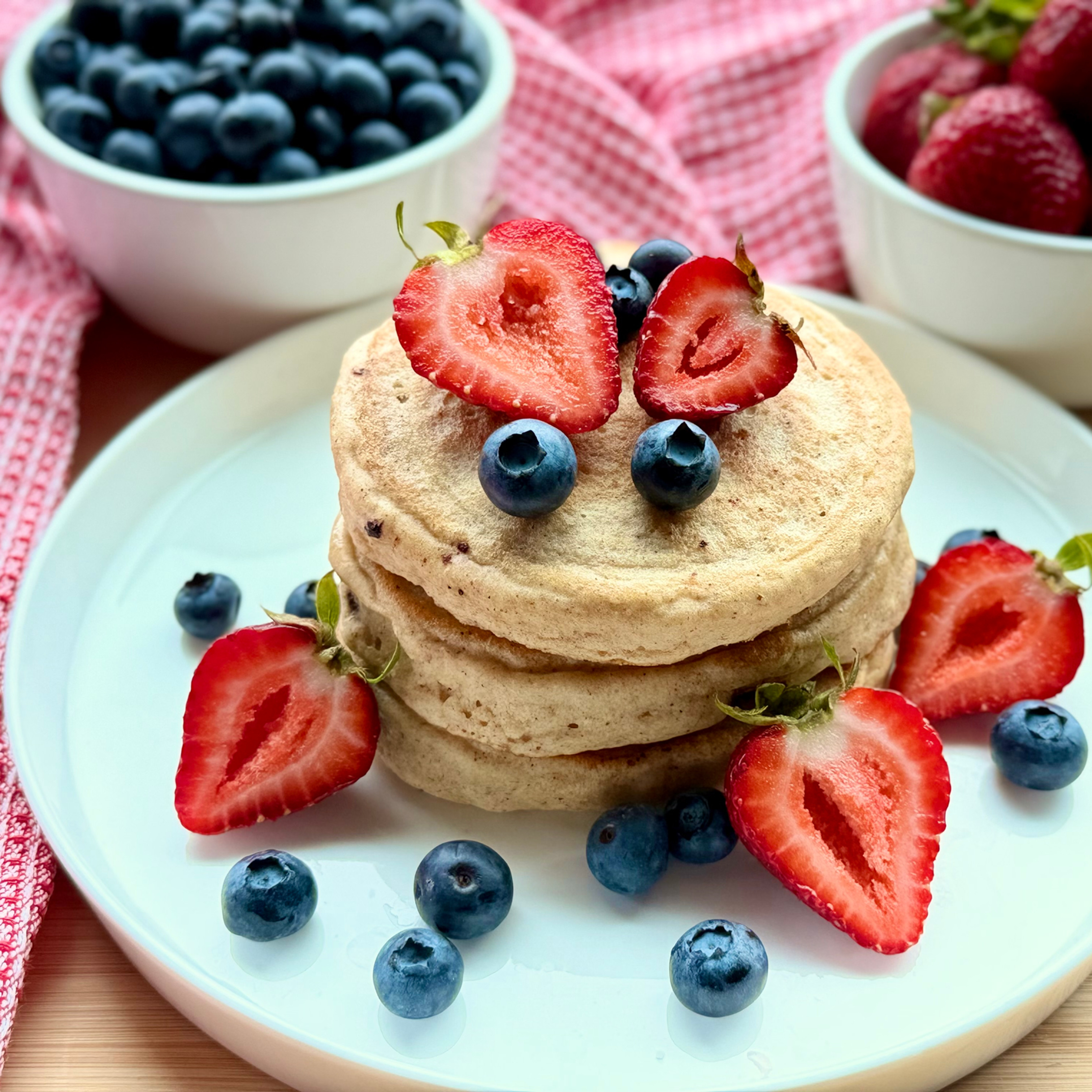 Stack of Fluffy Gluten-Free Pancakes topped with sliced strawberries and blueberries on a white plate, with bowls of berries and a pink cloth behind.
