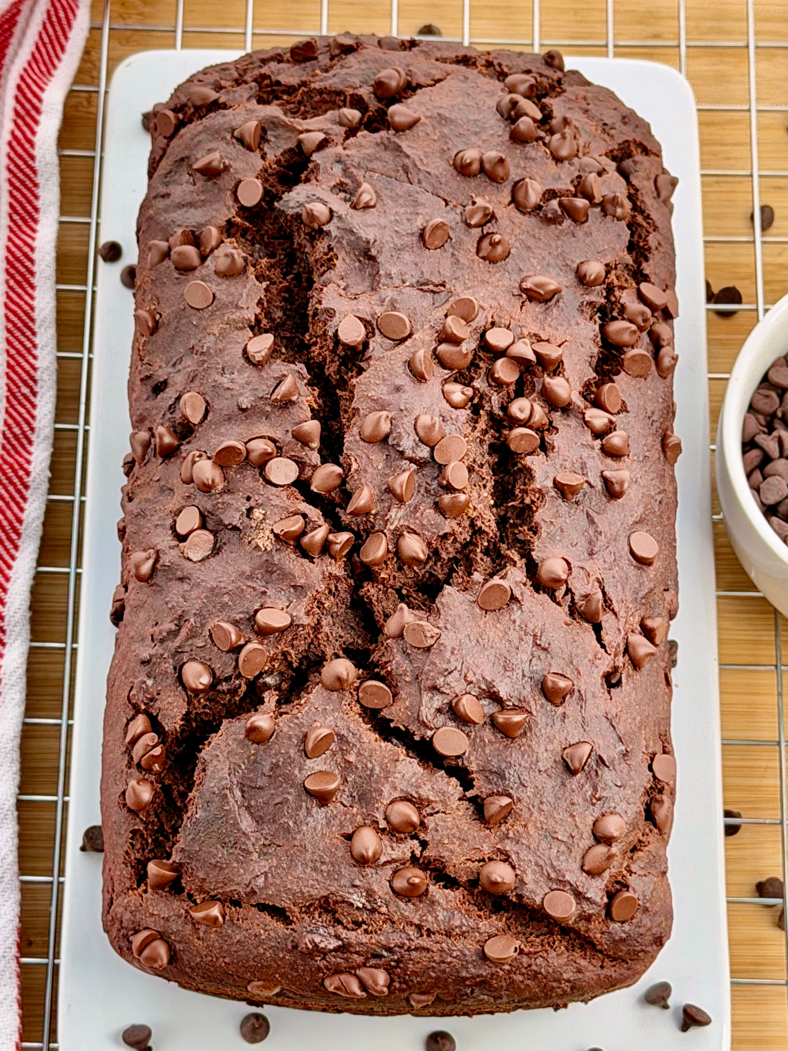 A Gluten-Free Double Chocolate Banana Bread load on a white platter, cooling on a rack beside a red-striped towel.