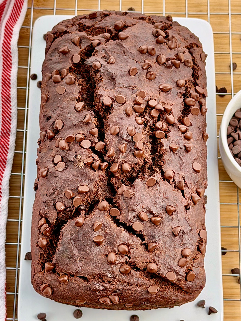 A Gluten-Free Double Chocolate Banana Bread load on a white platter, cooling on a rack beside a red-striped towel.