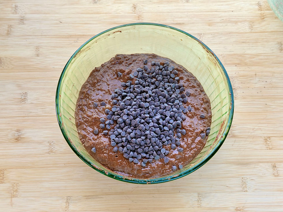 Glass bowl of Gluten-Free Double Chocolate Banana Bread batter topped with chocolate chips on a wooden cutting board, ready for baking