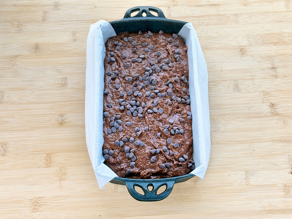 Gluten-Free Double Chocolate Banana Bread batter topped with chocolate chips in a parchment-lined loaf pan on a wooden cutting board.