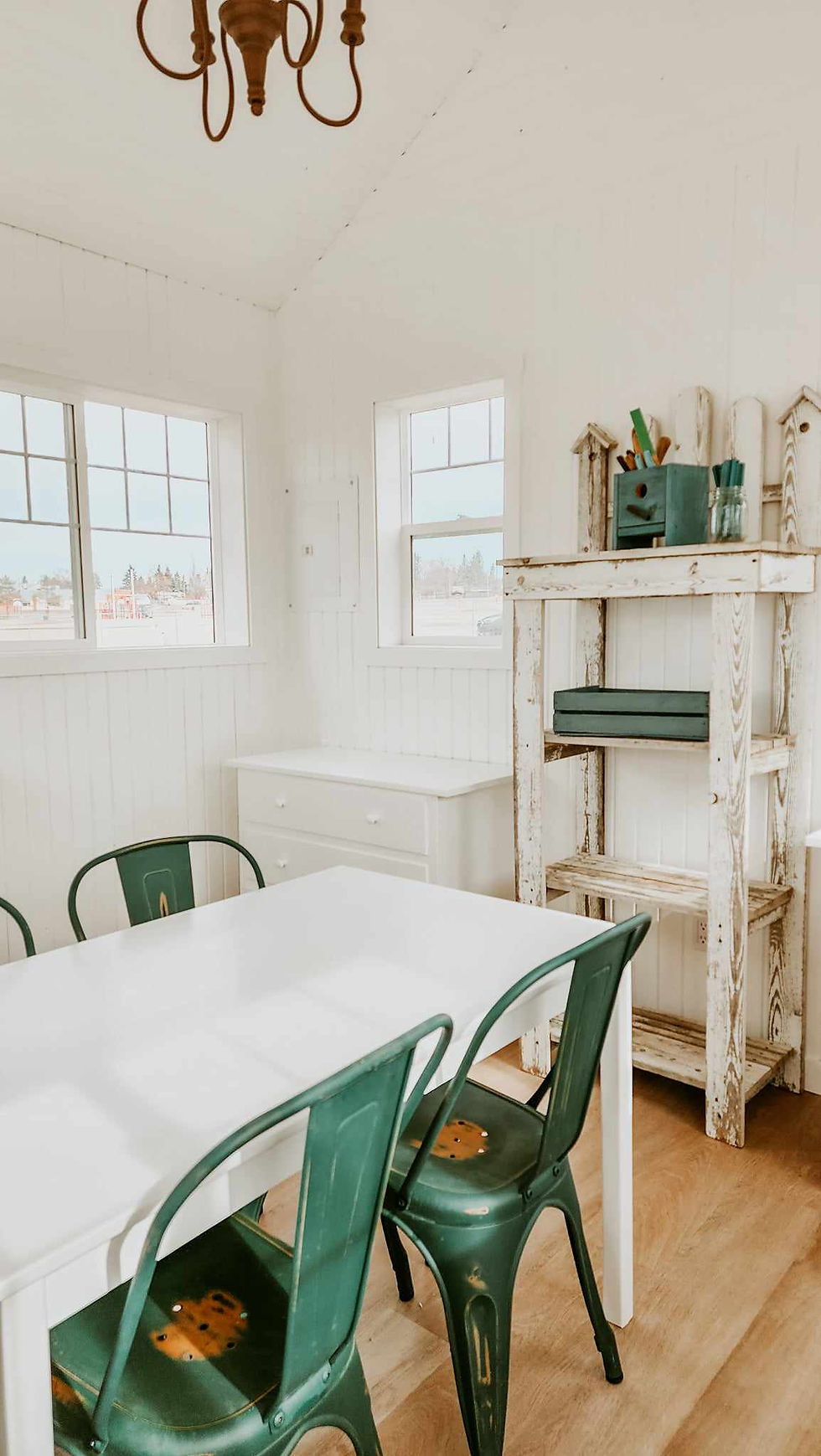 A white potting shed with green chairs and a long middle table. .