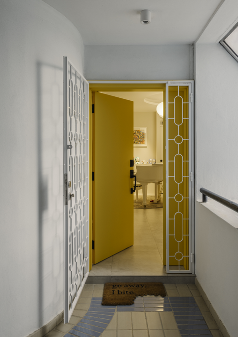 Entryway view through an open mustard-yellow door framed by a white patterned security gate, showing a glimpse of a brightly lit apartment interior with a white grand piano.