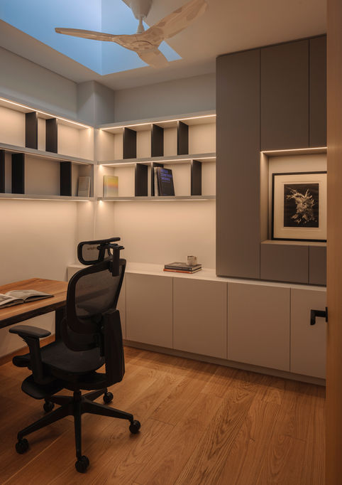 Modern home office interior with light wood flooring, a wooden desk with an ergonomic black office chair, built-in shelving with integrated warm lighting, and a skylight featuring an overhead ceiling fan.