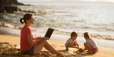 A woman works on her laptop at the beach while her children play, representing a healthy work-life balance and the freedom of modern work.