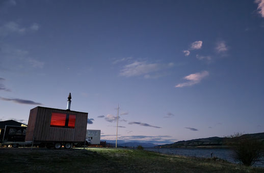 A mobile sauna trailer by Central Sauna with a glowing red window sits next to a lake at dusk in Cromwell, New Zealand