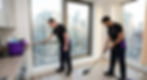Two professional male cleaners from 'Spify', wearing branded black and purple polo shirts, meticulously performing an end of tenancy clean in an empty modern apartment. One cleaner is detailed wiping the window sill, while the other vacuums the laminate floor. A large window reveals a bustling, dense city skyline outside, indicating an urban location.