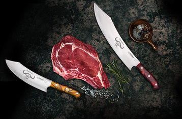 Ein saftiges rohes Steak liegt aufeiner dunklen Steinplatte, umringt von zwei scharfen Messern, Salz, Pfeffer und Rosmarin