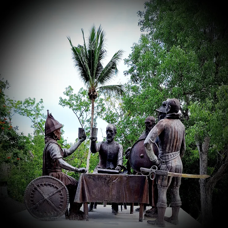 Bronze-colored monument from the Spanish colonial period. Two men clink glasses, they are sitting at a table, next to two soldiers.