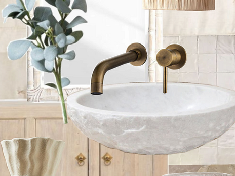 A tranquil organic modern bathroom by Kachi Interiors featuring a white carved stone vessel sink, antique bronze or brass wall-mounted faucet, and matching tap. Above the sink, a large mirror reflects a leafy green plant. To the left is a light wood vanity. A woven raffia wall sconce is on the right. In the foreground, a cream-colored vase with hand-painted floral designs sits next to a small, two-toned wooden stool. The floor is a light-colored marble.