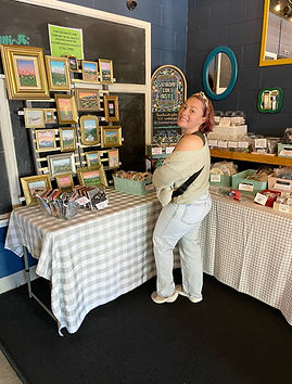 Smiling woman stands behind a table of art and crafts in a shop.