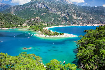 View of the Ölüdeniz lagoon, featuring its famous white sand beach and turquoise waters, surrounded by lush green mountains and a clear blue sky.