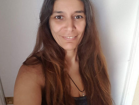 A woman with long brown hair smiles in a gray tank top. She stands in front of a white door, creating a casual and relaxed mood.