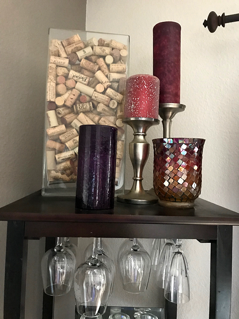 Red and purple candles, a mosaic candle holder, and a rectangular glass vase filled with wine cork on top of a wine rack holding wine glasses