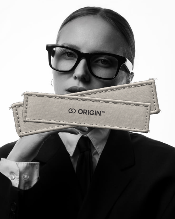 A black and white studio portrait shows a woman wearing glasses and a suit. Two fabric tags with the text "ORIGIN" are placed across her mouth.