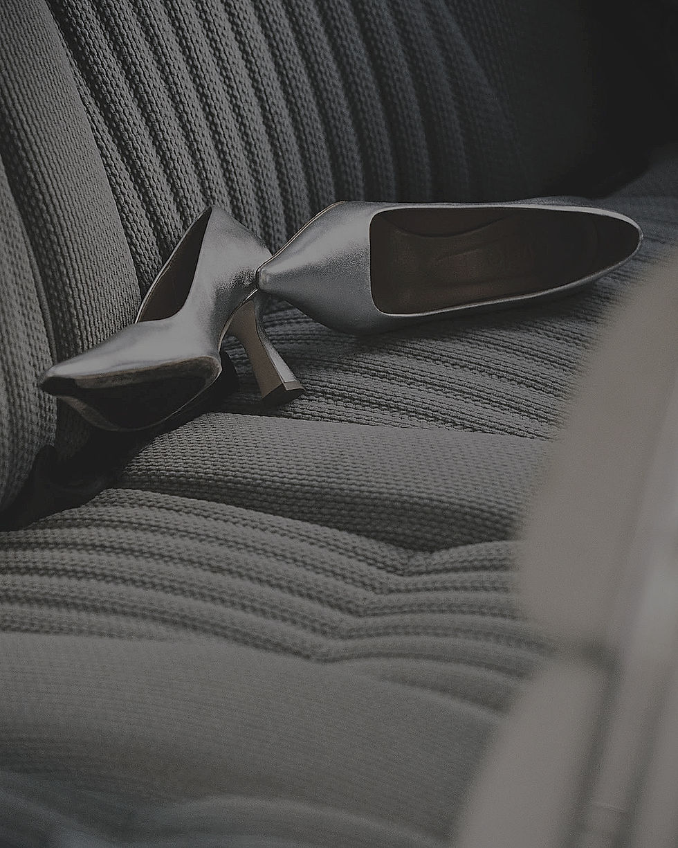 A pair of silver heels are placed on a textured gray surface. The shoes are positioned with their soles facing upwards, one slightly behind the other.