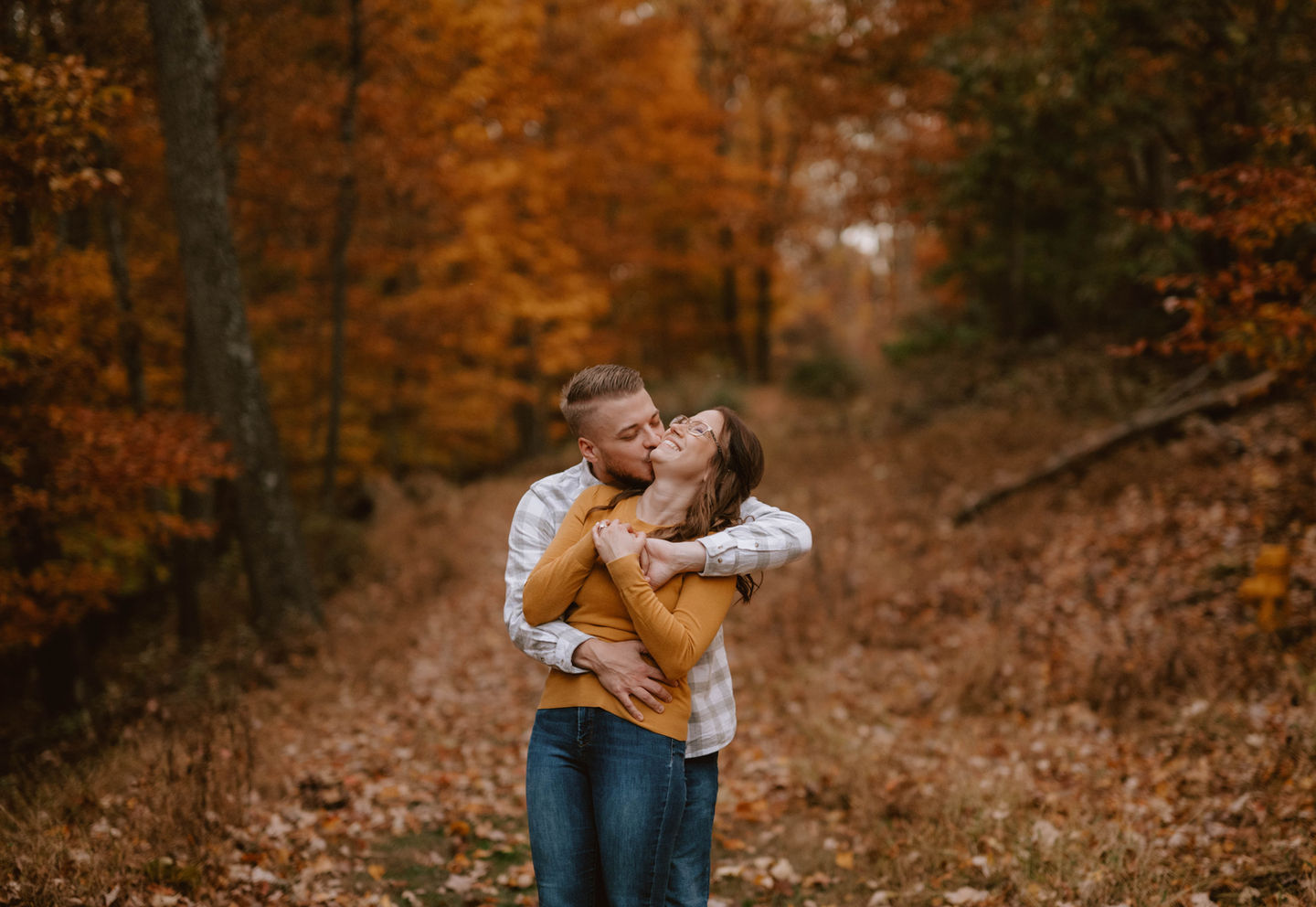A man is kissing a woman in an embrace while they stand in a forest clearing during autumn. The trees around them are filled with vibrant orange and yellow leaves, and fallen leaves cover the ground.