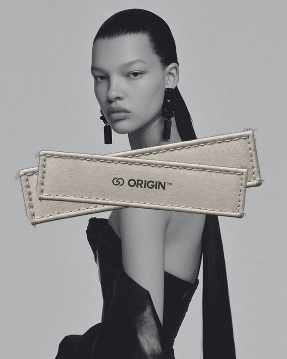 A black and white portrait of a woman wearing large earrings and a black garment. Two beige fabric tags with the "ORIGIN" logo are layered over the image.