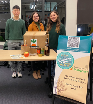 Photo of the three members of our team standing at a wood table that has a mock-up box on it with apples and jars in front of it. To the right of us is a mock-up of the physical signage on an A-Frame structure.