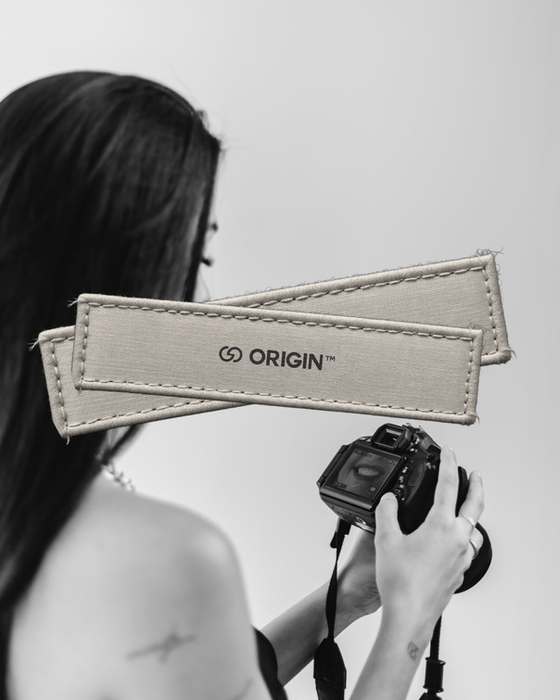 A woman is shown from behind holding a camera. Two fabric patches with the "ORIGIN" logo are layered over the image.