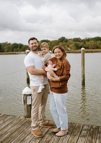A smiling family of four stands on a wooden dock by the water. The father holds a young boy, and the mother holds a newborn b