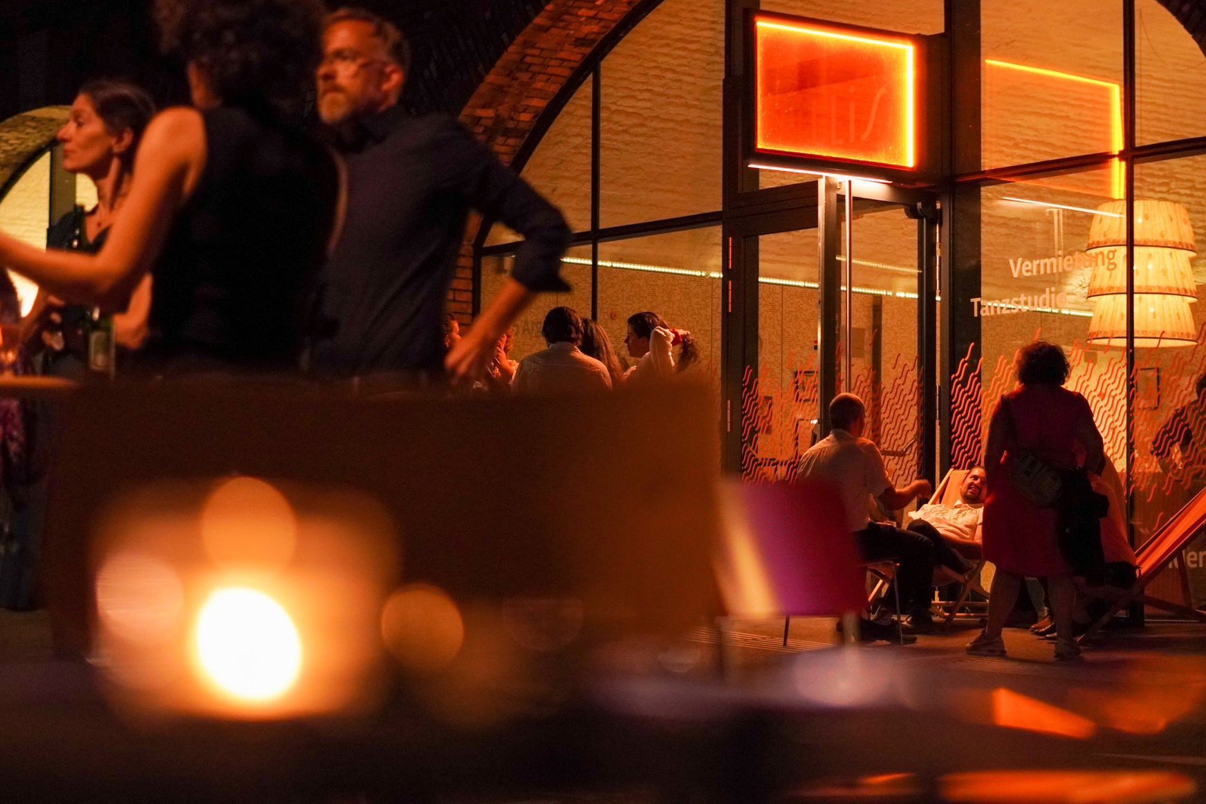 Stimmungsbild in orange, der Eingangsbogen von außen fotografiert, rundum stehen & sitzen Menschen und plaudern.