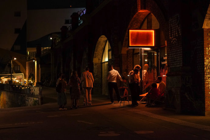 Abendansicht von außen auf Lillis Stadtbahnbögen, Leute stehen davor und unterhalten sich.