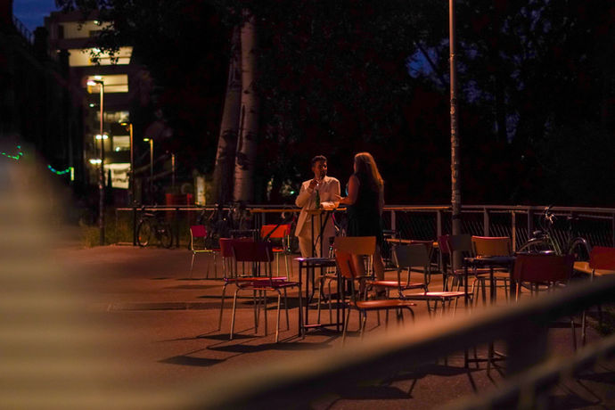 Aussicht von drinnen nach draußen, im Bogen sind Sessel aufgebaut und zwei Leute stehen mittendrin und unterhalten sich.