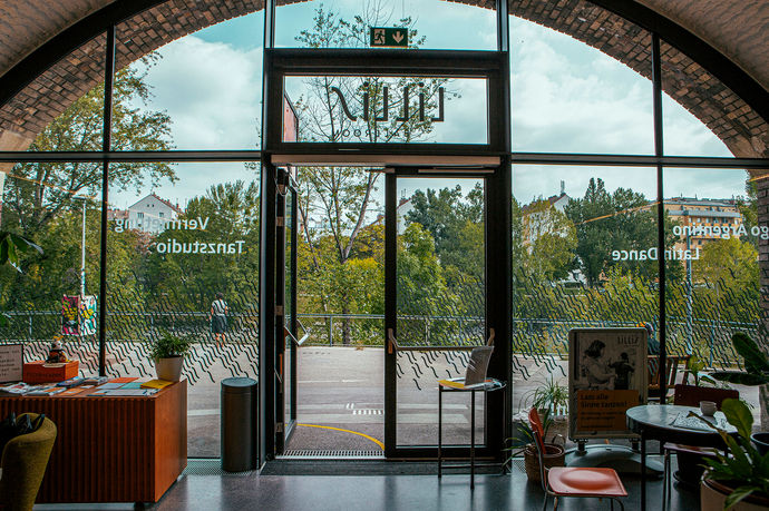 Eingang von innen fotografiert, Links ist ein Pult mit Flyer aufgebaut und rechts Tischchen mit Sesseln.