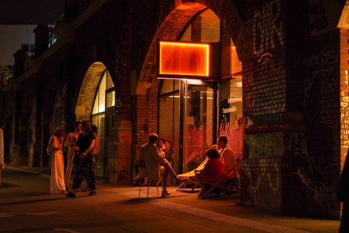 Lillis Stadtbahnbögen sind orange Beleuchtet, Leute sitzen und stehen davor und sind in Gespräche vertieft.