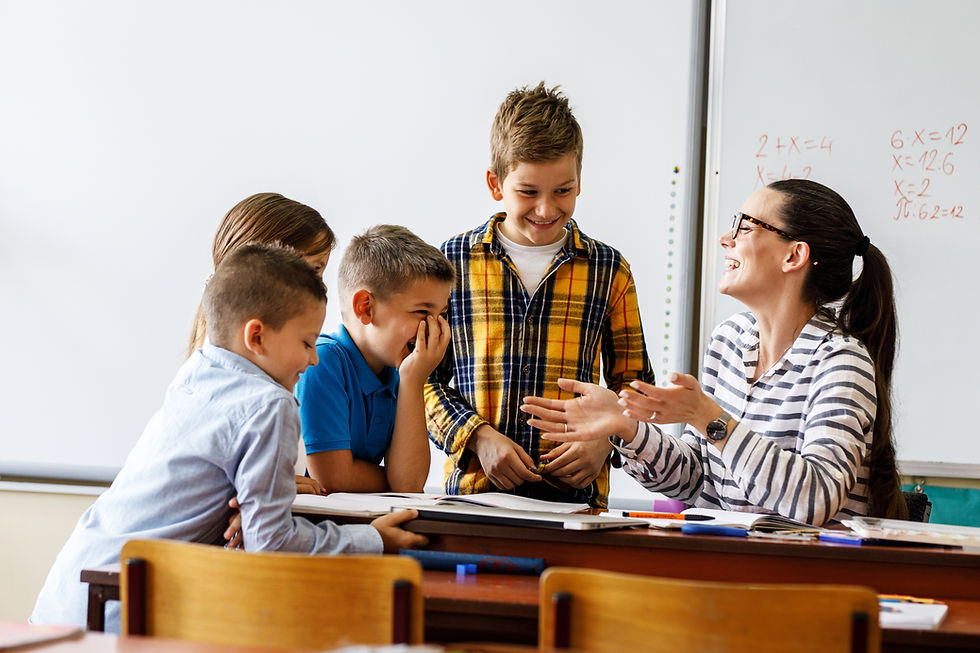Teacher and four kids laugh together in a classroom. The teacher points at the table. A whiteboard with math problems is visible behind them.