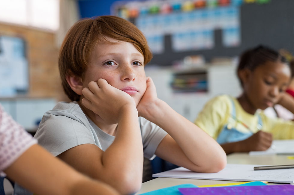 Child in a classroom, resting chin on hands, looks bored. Another child is writing. Blurred colorful background, with papers and pencils.