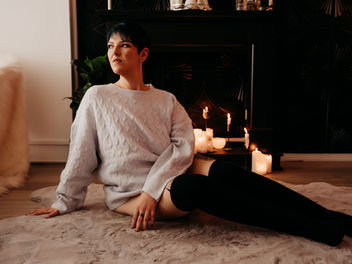 Ashley Stafford, intimate companion, in a cozy sweater and knee-high socks sitting on a fluffy rug with lit candles and dark decor in the background, creating a warm ambiance whilst gazing curiously over the other side of the room.
