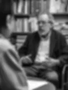Older attorney with glasses converses in an office filled with books. He gestures with hands, wearing a dark jacket. Pensive mood, black and white.