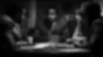 Four people in formal wear sit around a table in a dimly lit room, engaged in a serious discussion. Papers and a cup are on the table.