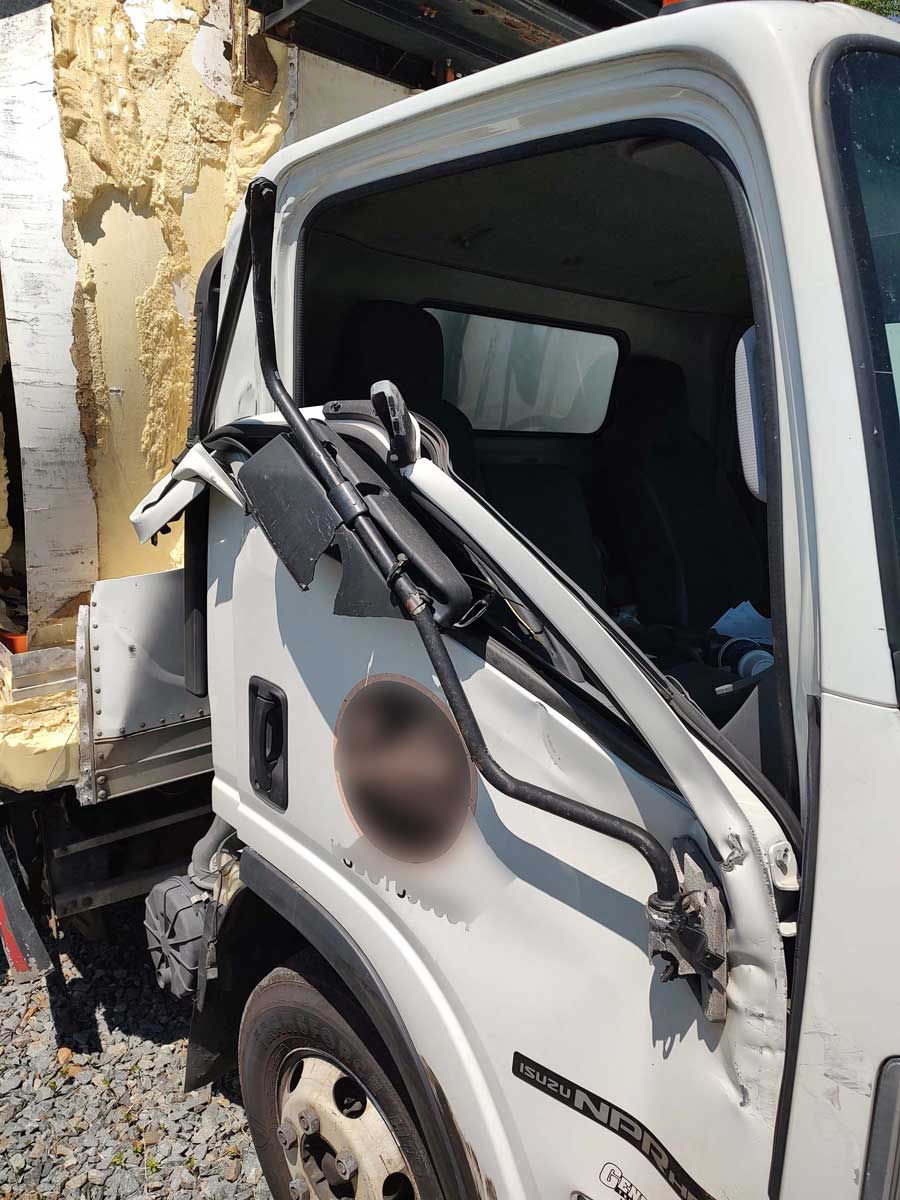 Damaged commercial truck with bent door and mirror, parked on gravel. Wall insulation visible in background. Bright sunlight highlights wreckage. Apis Law Personal Injury Attorney