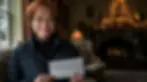 Woman in glasses holding a $280,000 check indoors with a lit Christmas tree in the background, smiling warmly. Dim, cozy setting.