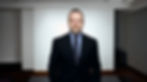 Attorney Keith F. Diaz in a dark suit stands in a modern, minimalistic room with a neutral background. He has a serious expression and a neat beard.