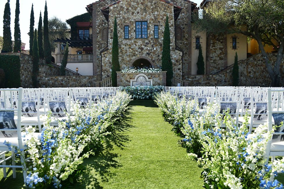 Outdoor wedding aisle lined with white chairs and blue-white flowers before a stone villa, calm sunny ceremony setup at Bella Collina.