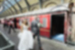 Bride in white and groom in gray suit walk smiling by red train in a station. Attendants in red vests stand nearby. Bright, joyous mood.