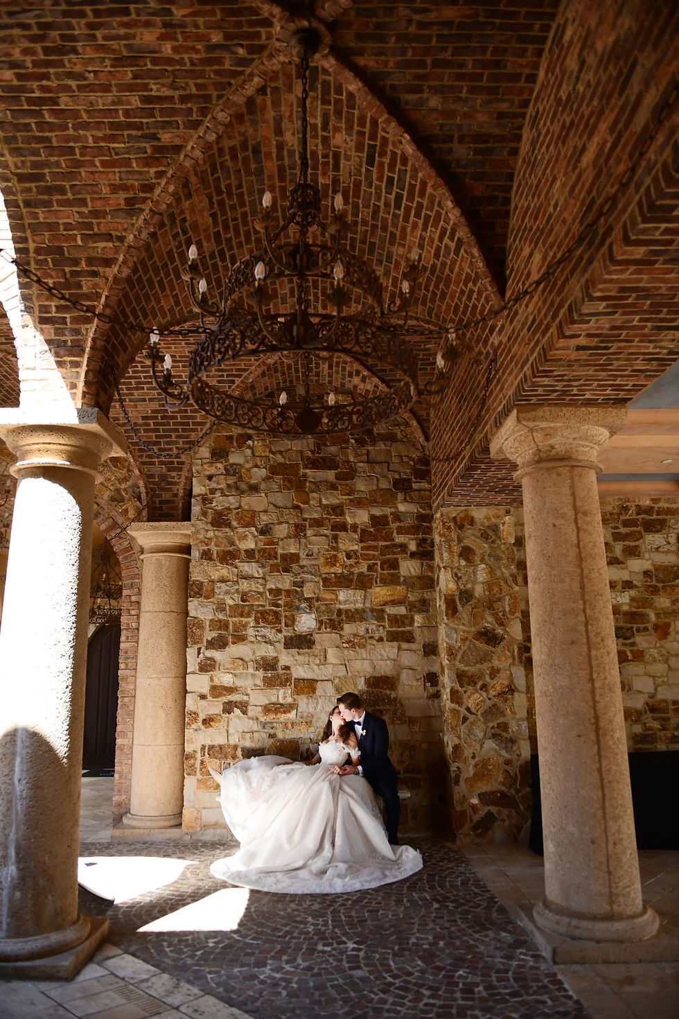 Bride and groom kiss in a sunlit stone courtyard beneath iron chandeliers and brick arches, creating a romantic mood at Bella Collina.