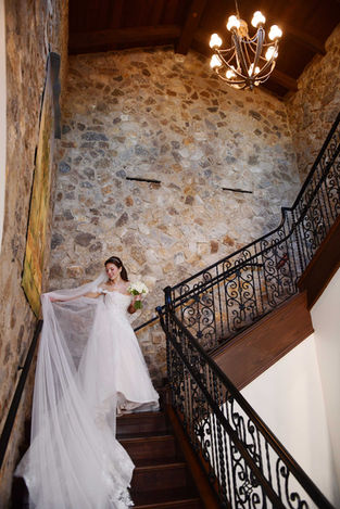 Elegant bridal portrait on a grand stone staircase featuring a bride in a sparkling white wedding gown with a flowing cathedral veil, captured in a luxurious rustic wedding venue with warm chandelier lighting and timeless romantic architecture.
