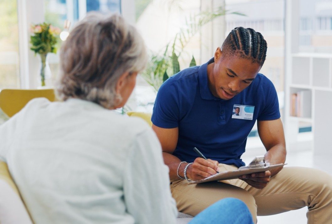 A male community health worker taking notes while talking with an older woman
