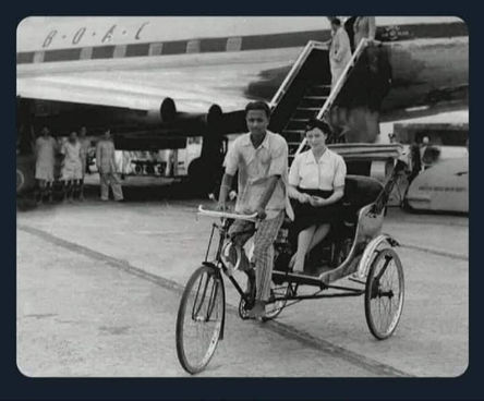 “An air hostess in a skirt and uniform sits in a hand-pulled rickshaw outside Calcutta airport, poised for duty amid the morning bustle.”
