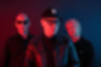 Three men pose under red and blue studio lighting; center wears an N cap and glasses, all with serious expressions.