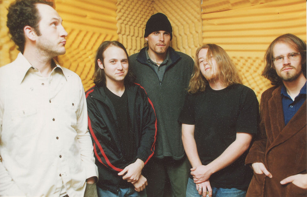 Five men pose in a yellow acoustic booth, wearing casual clothes and serious expressions in a band-style portrait.