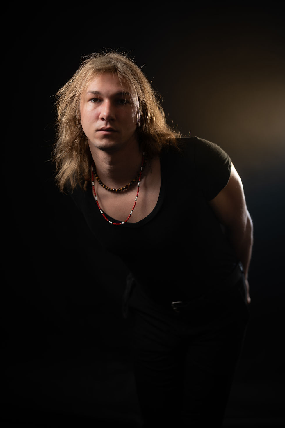 Studio portrait of a young person with long blond hair, black shirt, and beaded necklaces against a dark background, serious expression.