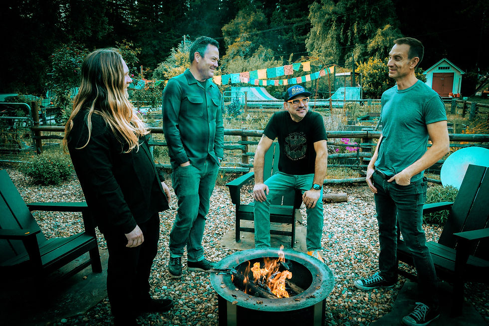 Four adults chat and smile around a lit fire pit in a rustic garden, with colorful prayer flags and trees in the background.
