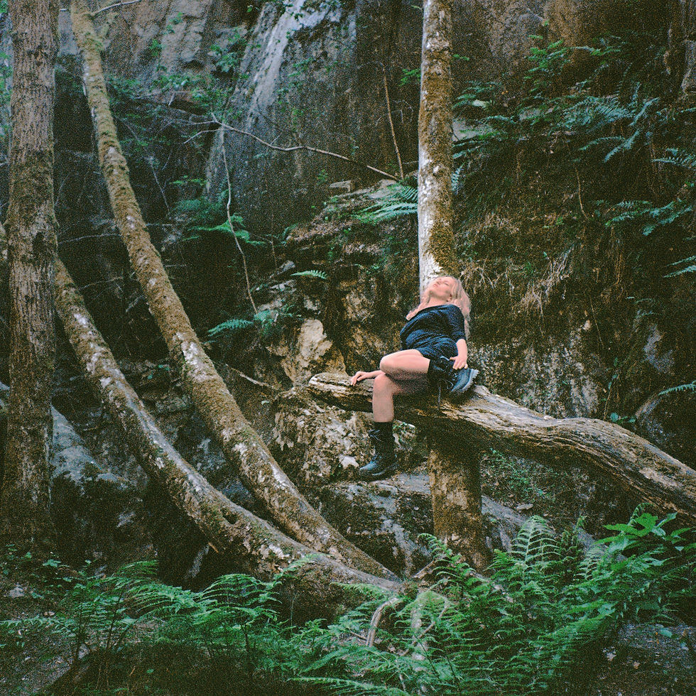 Woman in a dark outfit reclines on a fallen tree in a lush green forest, surrounded by ferns and mossy trunks.